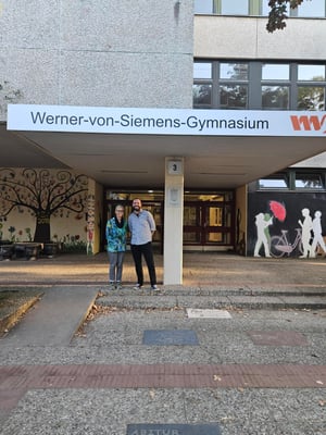 An image for Ms. Rosa Spyra and Sebastian Telschow, smiling and standing outside the main entrance of Werner von Siemens Gymnasium